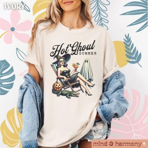 Pu&ograve; includere: T-shirt color avorio con la scritta "Hot Ghoul Summer" e una grafica di una strega che si rilassa su una sedia a sdraio con un cocktail, una zucca, un fantasma e un alligatore. La maglietta ha le maniche arrotolate.