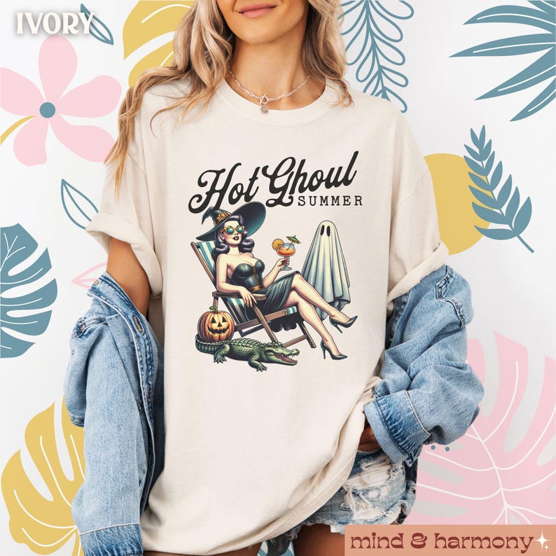 Pu&ograve; includere: T-shirt color avorio con la scritta "Hot Ghoul Summer" e una grafica di una strega che si rilassa su una sedia a sdraio con un cocktail, una zucca, un fantasma e un alligatore. La maglietta ha le maniche arrotolate.