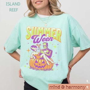 May include: A light teal t-shirt with the words "SUMMER Ween" in a retro font. The graphic features Frankenstein and his bride sitting on a jack-o'-lantern, holding cocktails. The shirt has a relaxed fit and the words "ISLAND REEF" are printed at the top.