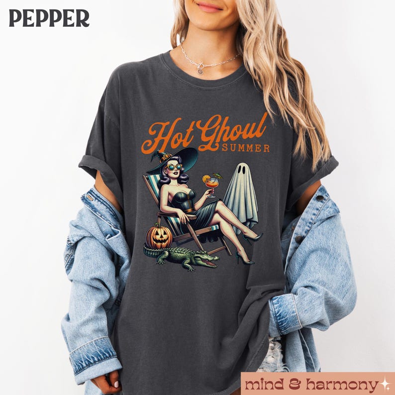 Pu&ograve; includere: T-shirt grigio scuro con la scritta "Hot Ghoul Summer" in arancione. La grafica presenta una donna con cappello da strega, occhiali da sole e abito nero, che si rilassa su una sedia a sdraio con un cocktail, una zucca, un fantasma e un alligatore.