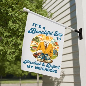 May include: White flag with the text "IT'S A Beautiful Day TO Protect & Defend MY NEIGHBORS" in blue script. The flag features a cartoon illustration of a yellow sweater, houses, a train, and blue sneakers.