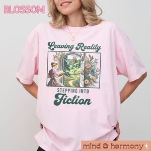 May include: Pink t-shirt with a graphic of a woman reading a book in front of a doorway with the text "Leaving Reality Stepping Into Fiction".