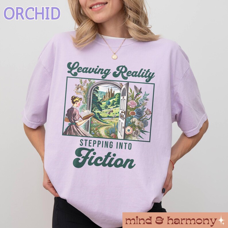 May include: A lavender t-shirt with the text "Leaving Reality Stepping Into Fiction" in green lettering. The design features a woman reading a book in front of a doorway leading to a fantasy landscape.