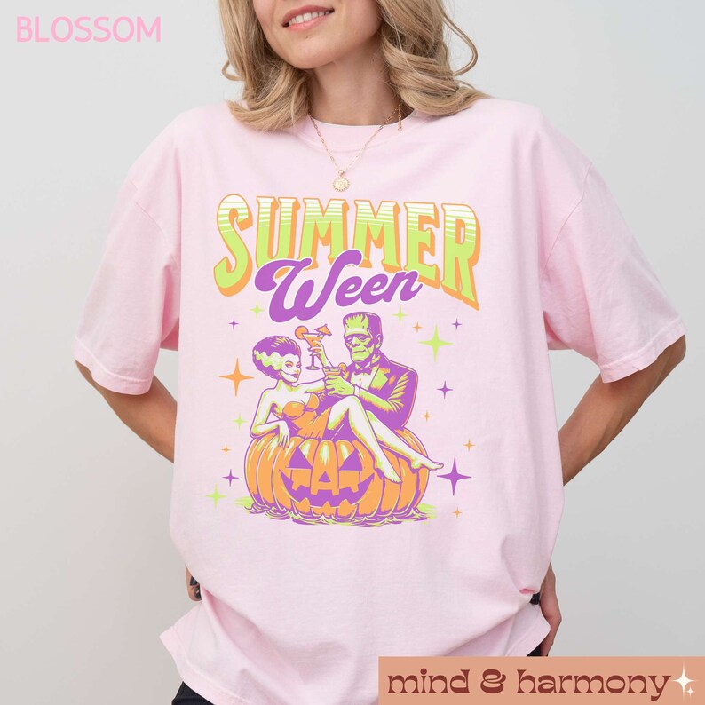 May include: Light pink t-shirt with the text "SUMMER WEEN" in green and purple. The graphic features a cartoon couple, a bride and Frankenstein, sitting on a jack-o'-lantern, holding cocktails. The shirt has short sleeves.