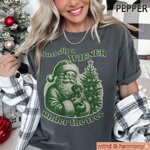 May include: A dark grey t-shirt with a green illustration of Santa Claus holding a dachshund under a Christmas tree. The text "Just slip a WIENER under the tree" is printed on the shirt.