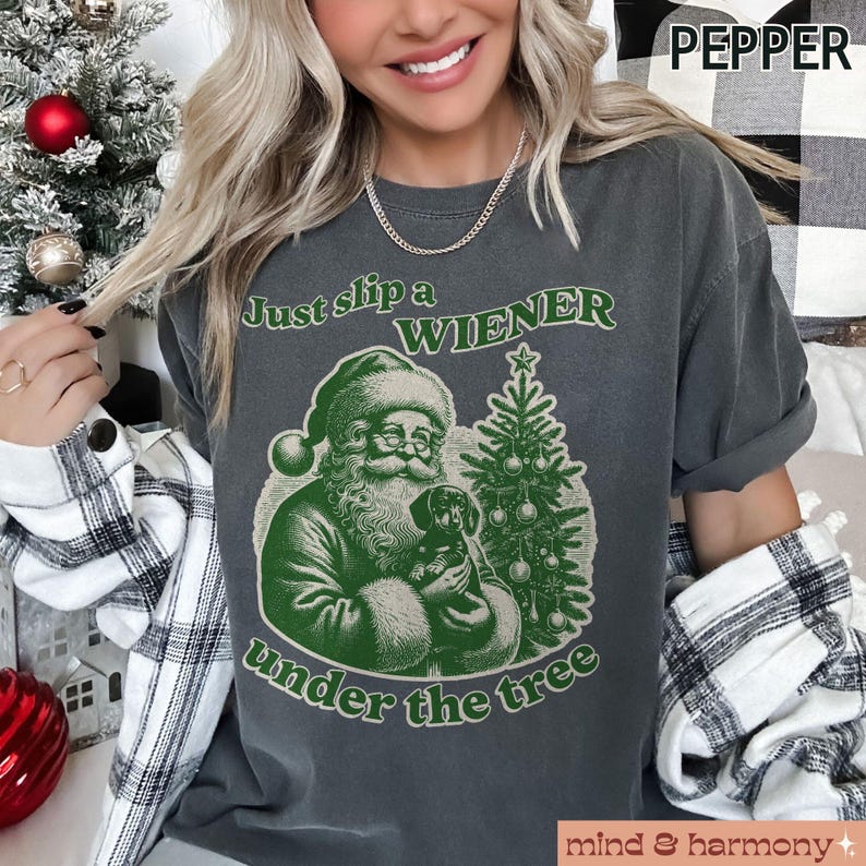 May include: A dark grey t-shirt with a green illustration of Santa Claus holding a dachshund under a Christmas tree. The text "Just slip a WIENER under the tree" is printed on the shirt.