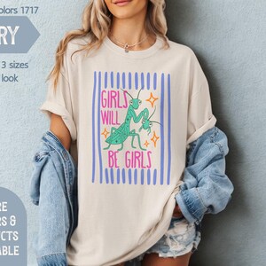 Puede incluir: Camiseta de color marfil con el texto "GIRLS WILL BE GIRLS" en rosa y turquesa, con un gr&aacute;fico de dos mantis religiosas. La camiseta tiene rayas verticales azules y forma parte de la colecci&oacute;n "Mind & Harmony Gifts".