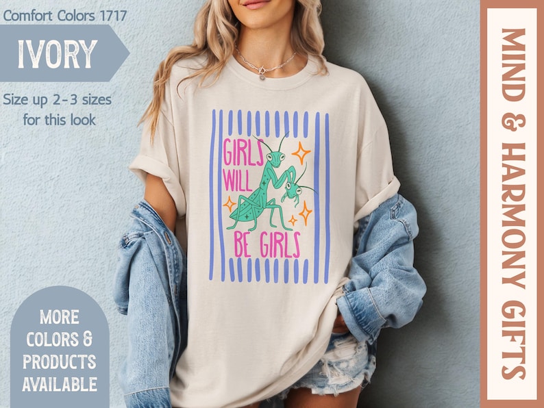 Puede incluir: Camiseta de color marfil con el texto "GIRLS WILL BE GIRLS" en rosa y turquesa, con un gr&aacute;fico de dos mantis religiosas. La camiseta tiene rayas verticales azules y forma parte de la colecci&oacute;n "Mind & Harmony Gifts".