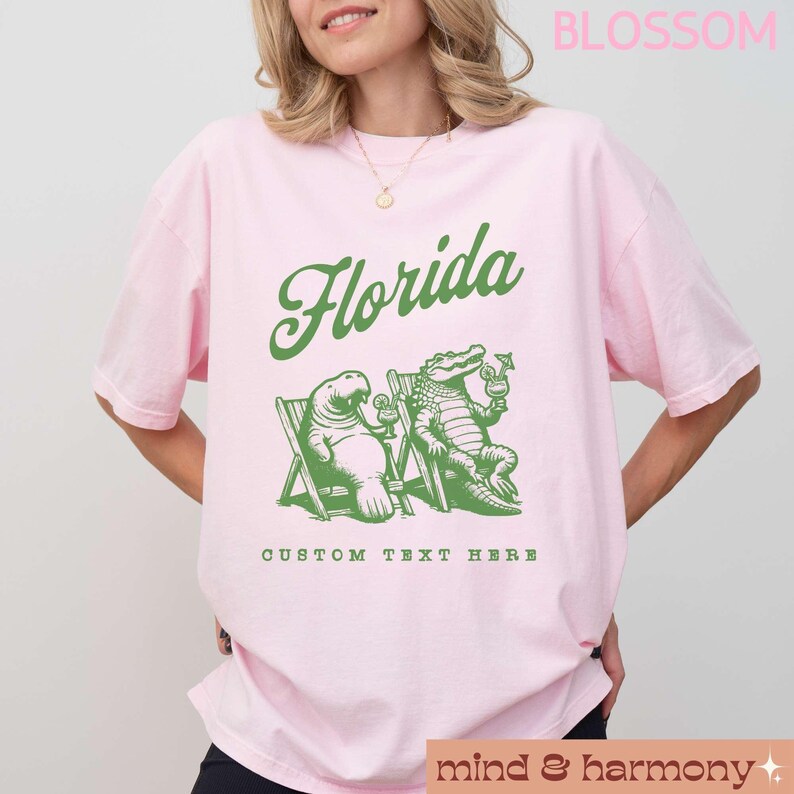 May include: Pink t-shirt with a green graphic of a manatee and alligator sitting in lounge chairs with drinks. The text "Florida" is above the graphic and "Custom Text Here" is below the graphic.