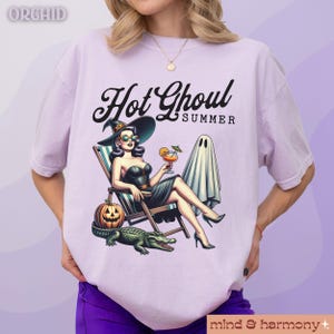 Pu&ograve; includere: T-shirt lilla chiaro con una grafica di una donna con cappello da strega, occhiali da sole e un vestito nero, che si rilassa su una sedia a sdraio con un cocktail. Il testo "Hot Ghoul Summer" &egrave; sopra l'immagine. Una zucca, un alligatore e un fantasma sono anche nell'immagine.