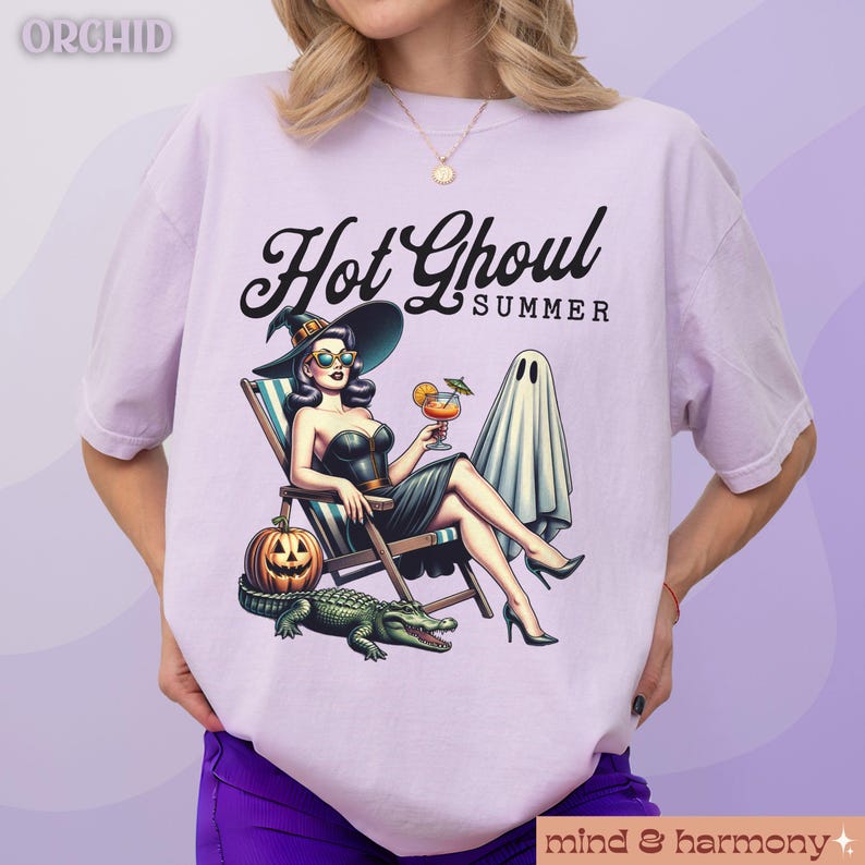 Pu&ograve; includere: T-shirt lilla chiaro con una grafica di una donna con cappello da strega, occhiali da sole e un vestito nero, che si rilassa su una sedia a sdraio con un cocktail. Il testo "Hot Ghoul Summer" &egrave; sopra l'immagine. Una zucca, un alligatore e un fantasma sono anche nell'immagine.