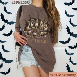 Peut inclure: Un t-shirt marron avec un motif de quatre sorci&egrave;res assises autour d'une table avec des cr&acirc;nes et des os. Le texte "Girls will be girls" est imprim&eacute; sur le t-shirt.