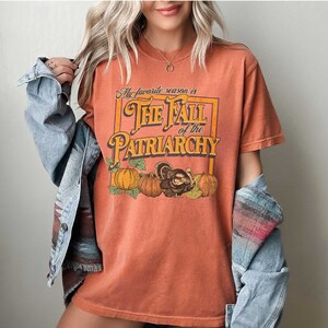 May include: A burnt orange t-shirt featuring a graphic print of a vintage-style box with the text "My favourite season is The Fall of the Patriarchy" and a turkey and pumpkins.