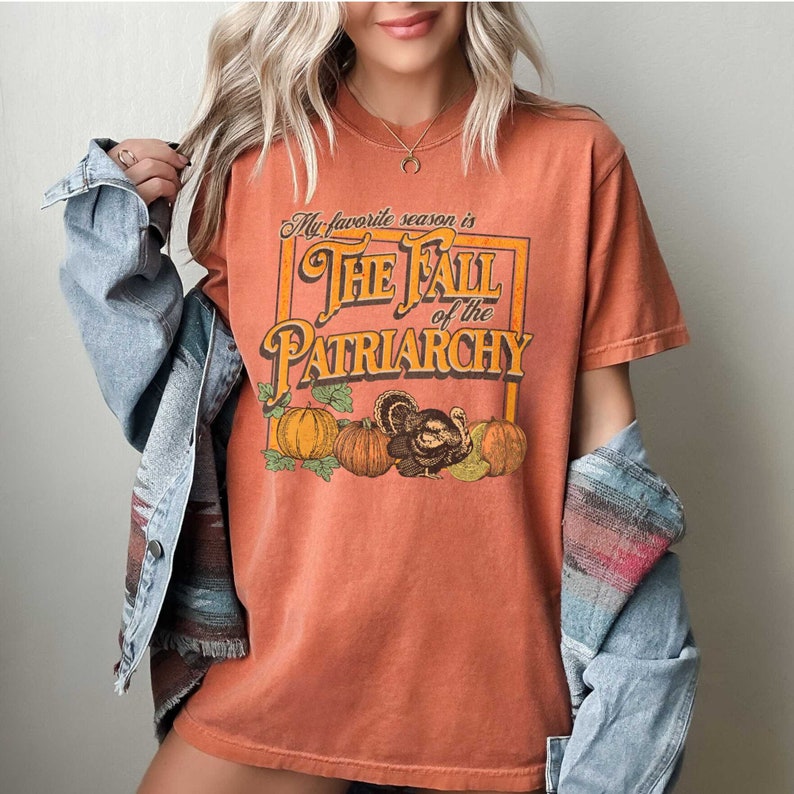 May include: A burnt orange t-shirt featuring a graphic print of a vintage-style box with the text "My favourite season is The Fall of the Patriarchy" and a turkey and pumpkins.