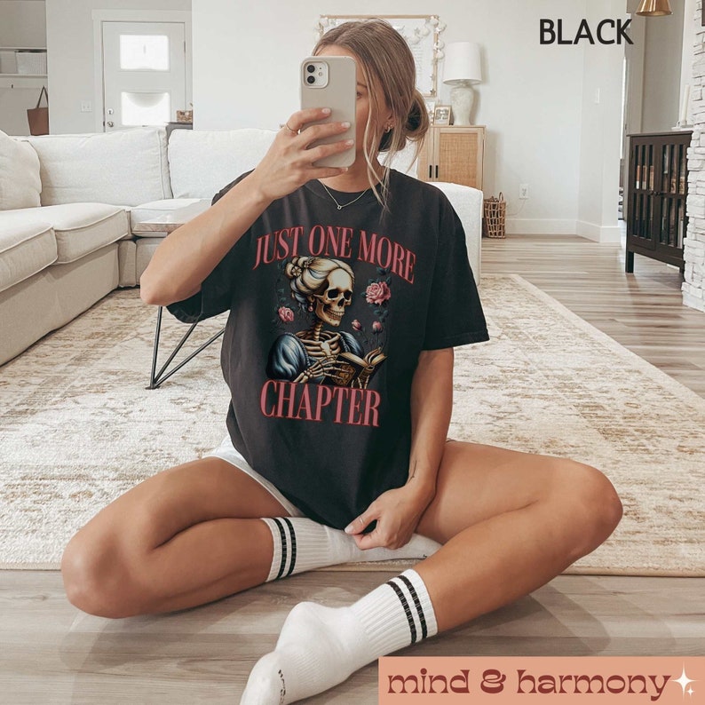 May include: A black t-shirt with a graphic of a skeleton woman reading a book with the text "Just One More Chapter".