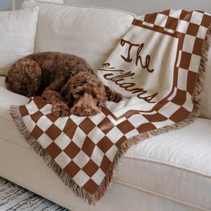 May include: A brown and white checkered throw blanket with the name "The Williams" in brown script is draped over a white sofa. A brown dog is resting on the blanket. The blanket has a fringed edge.