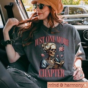 May include: A gray t-shirt with the text "Just One More Chapter" printed in pink. A skeleton wearing a dress and holding a book is illustrated below the text. The skeleton is surrounded by pink flowers.
