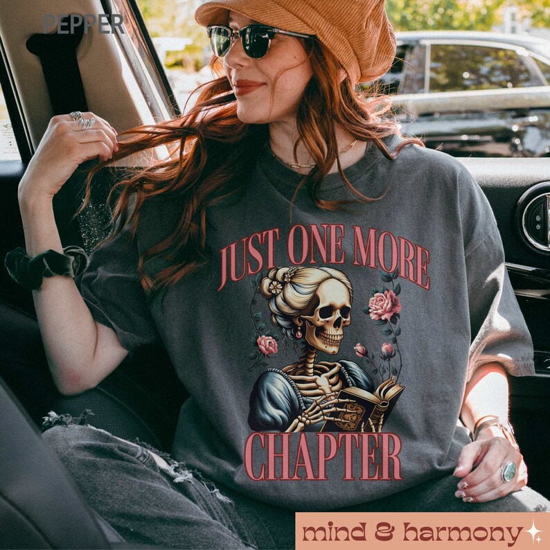 May include: A gray t-shirt with the text "Just One More Chapter" printed in pink. A skeleton wearing a dress and holding a book is illustrated below the text. The skeleton is surrounded by pink flowers.