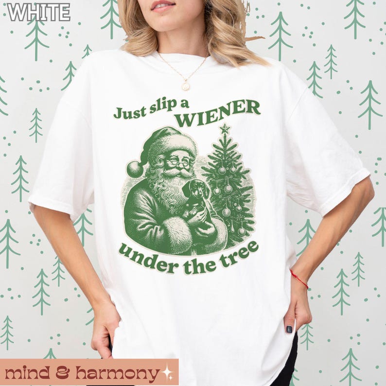 May include: White t-shirt featuring a green illustration of Santa Claus holding a dachshund under a Christmas tree. The text "Just slip a WIENER under the tree" is printed on the shirt.