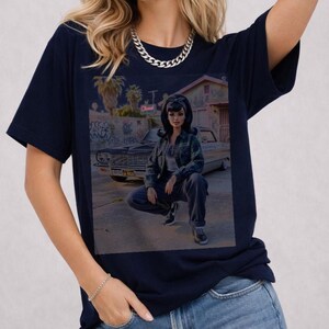May include: Navy blue t-shirt featuring a graphic of a doll in front of a classic car. The doll is wearing a plaid shirt and jeans. The background includes graffiti, palm trees, and a building. A silver chain necklace is visible.
