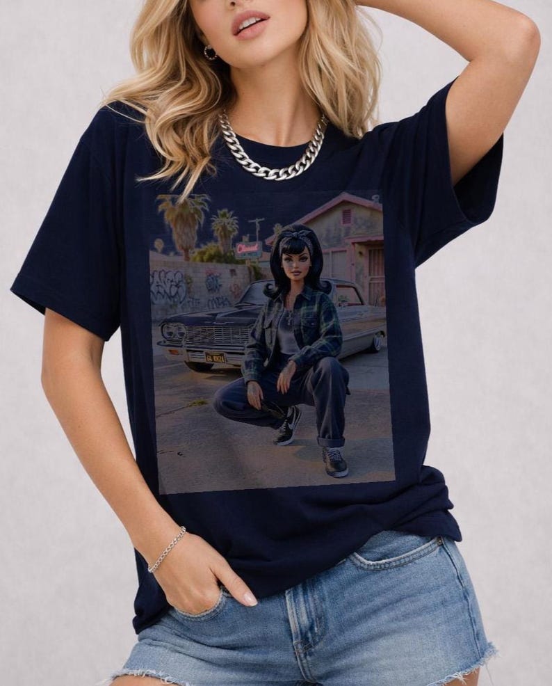 May include: Navy blue t-shirt featuring a graphic of a doll in front of a classic car. The doll is wearing a plaid shirt and jeans. The background includes graffiti, palm trees, and a building. A silver chain necklace is visible.