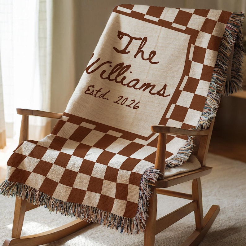 May include: A brown and cream-colored throw blanket with a checkerboard pattern and fringe detail is draped over a wooden rocking chair. The blanket features the text "The Williams Estd. 2026" in a brown script font.