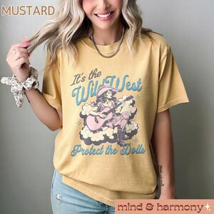 Może przedstawiać: Ż&oacute;łta koszulka w kolorze musztardowym z grafiką w stylu vintage przedstawiającą kobietę grającą na gitarze z tekstem "It's the Wild West Protect the Dolls".