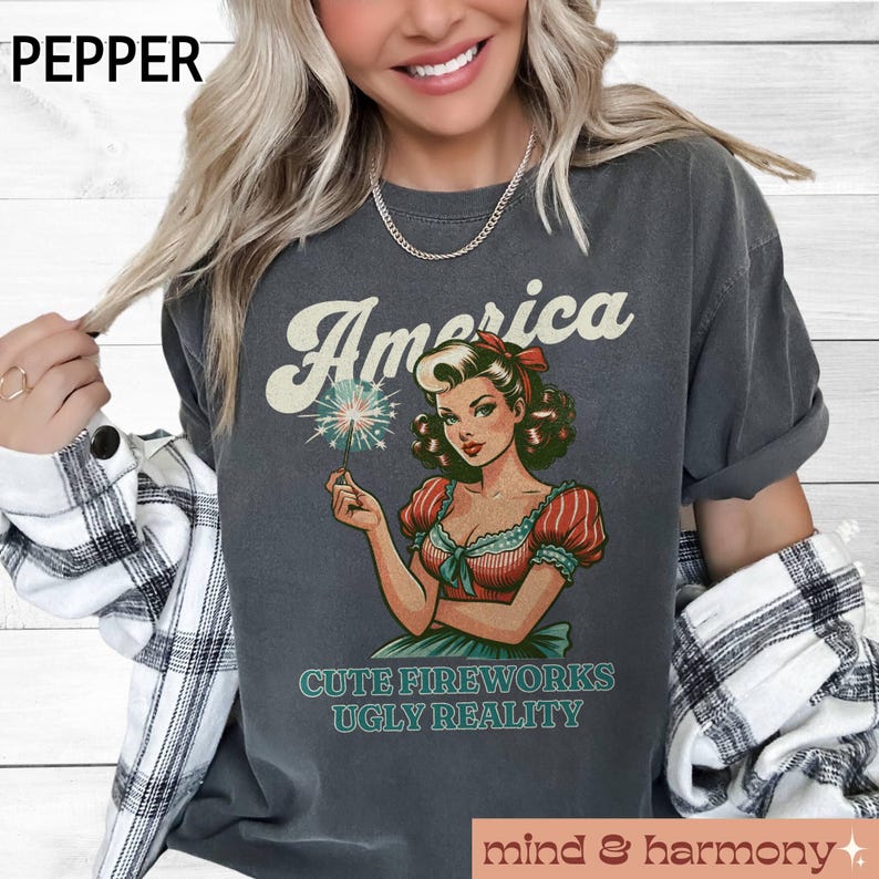 May include: Dark gray t-shirt with the word "America" above a vintage-style illustration of a woman holding a sparkler. The text "CUTE FIREWORKS UGLY REALITY" is below the illustration. The shirt has rolled-up sleeves.