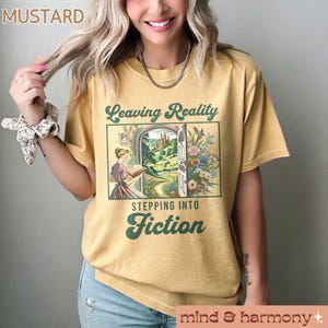 May include: A mustard yellow t-shirt with a vintage-style graphic that says "Leaving Reality Stepping Into Fiction". The graphic features a woman reading a book in front of a door that opens to a magical forest.