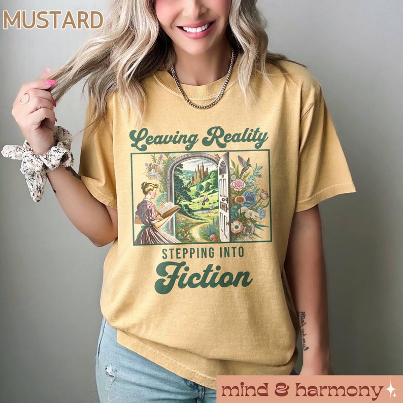 May include: A mustard yellow t-shirt with a vintage-style graphic that says "Leaving Reality Stepping Into Fiction". The graphic features a woman reading a book in front of a door that opens to a magical forest.