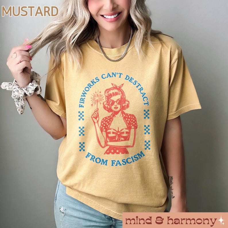 May include: Mustard-coloured t-shirt featuring a retro graphic of a woman holding a firework. The text reads "Fireworks Can't Distract From Fascism" in a blue, circular design. The shirt has short sleeves and a relaxed fit.