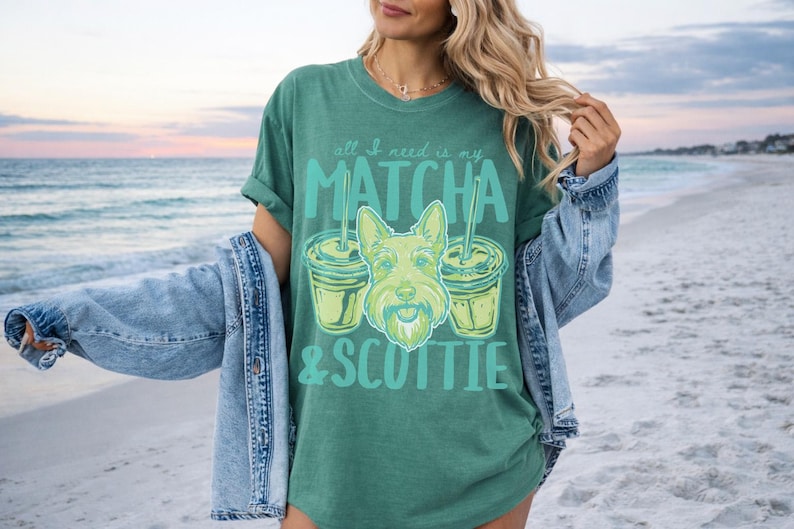 May include: Green t-shirt with the text "all I need is my MATCHA & SCOTTIE" in teal. The graphic features a Scottie dog and two matcha drinks. The shirt is worn with a denim jacket on a beach.