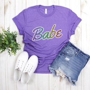 May include: Purple t-shirt with a rainbow leopard print "Babe" graphic.