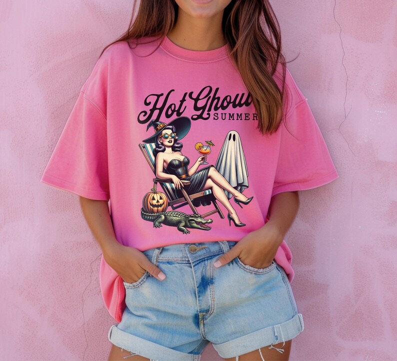 Pu&ograve; includere: Maglietta rosa con una grafica di una donna con un cappello da strega che si rilassa su una sedia a sdraio, con in mano un cocktail. Il testo "Hot Ghoul Summer" &egrave; sopra l'immagine. Una zucca, un alligatore e un fantasma sono anche nell'immagine.
