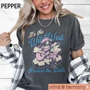 Może przedstawiać: Szary t-shirt z grafiką w stylu vintage przedstawiającą kobietę grającą na gitarze z tekstem "It's the Wild West Protect the Dolls".