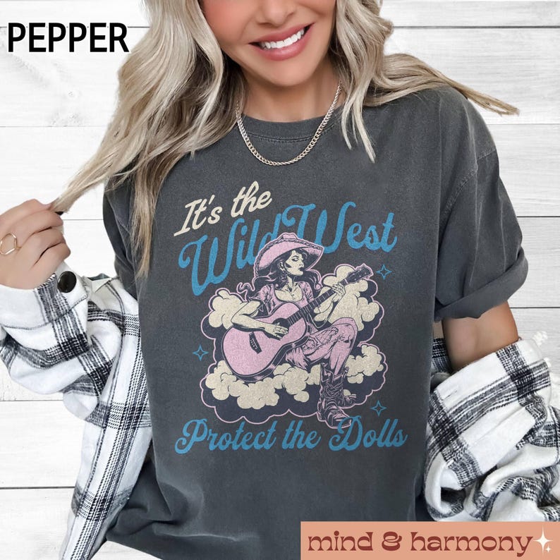 Może przedstawiać: Szary t-shirt z grafiką w stylu vintage przedstawiającą kobietę grającą na gitarze z tekstem "It's the Wild West Protect the Dolls".