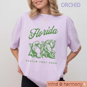 May include: A lavender t-shirt with the word "Florida" in green text. The shirt features a cartoon manatee and alligator sitting on beach chairs with drinks. The text "Custom Text Here" is below the image.