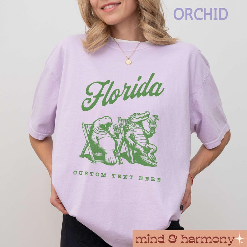 May include: A lavender t-shirt with the word "Florida" in green text. The shirt features a cartoon manatee and alligator sitting on beach chairs with drinks. The text "Custom Text Here" is below the image.