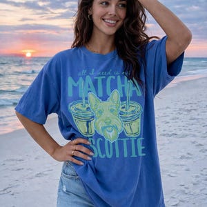May include: Blue t-shirt with the text "all I need is matcha scottie" and a graphic of a Scottish Terrier dog and two matcha drinks. The shirt is worn on a beach at sunset.