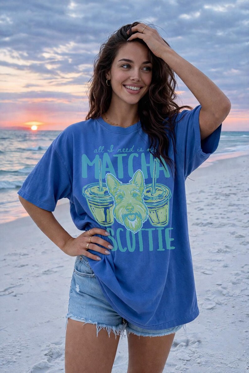 May include: Blue t-shirt with the text "all I need is matcha scottie" and a graphic of a Scottish Terrier dog and two matcha drinks. The shirt is worn on a beach at sunset.