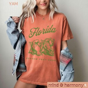 May include: A burnt orange t-shirt with a green graphic of a manatee and alligator sitting in beach chairs with drinks. The text "Florida" is in a green cursive font above the graphic. The text "CUSTOM TEXT HERE" is below the graphic.