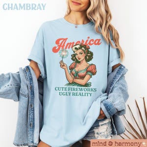 May include: Light blue t-shirt with the word "America" in red script above a vintage-style illustration of a woman holding a sparkler. The text "CUTE FIREWORKS UGLY REALITY" is below the illustration. The shirt is made of a soft material.