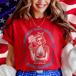 May include: A red t-shirt with a vintage-style illustration of a woman holding a firework. The text around the image reads "Fireworks Can't Distract From Fascism" in blue. The woman is wearing sunglasses and a patriotic outfit.