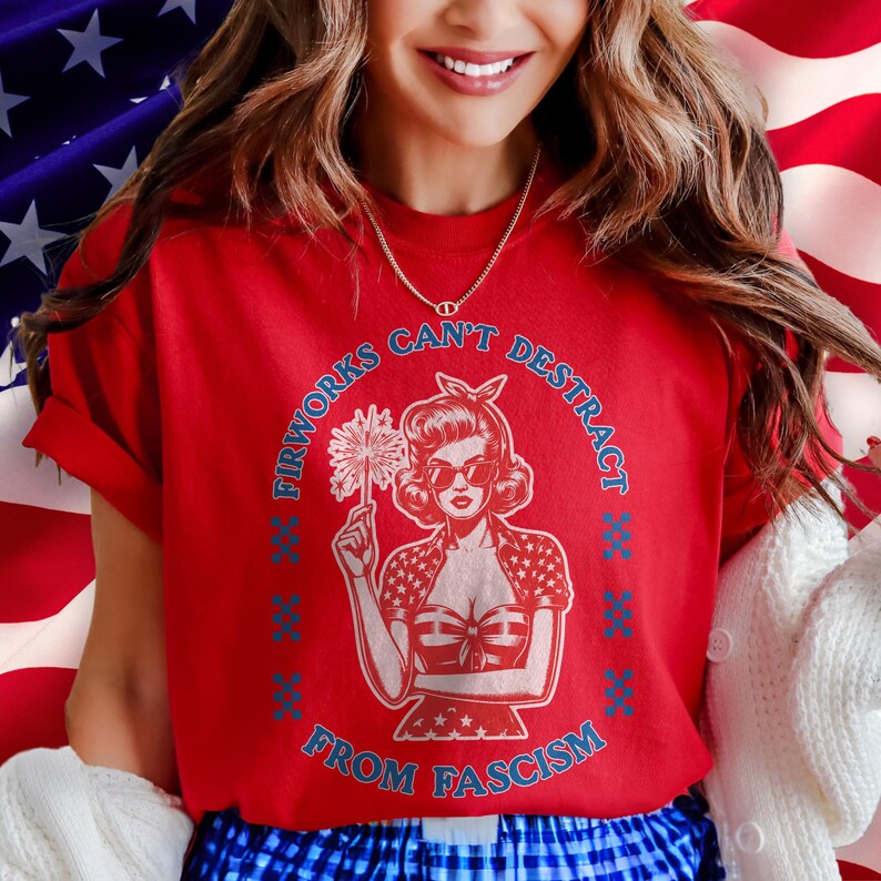 May include: A red t-shirt with a vintage-style illustration of a woman holding a firework. The text around the image reads "Fireworks Can't Distract From Fascism" in blue. The woman is wearing sunglasses and a patriotic outfit.