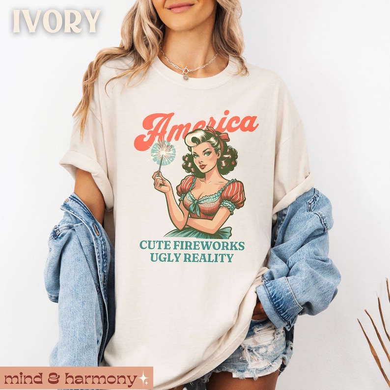 May include: Ivory-colored t-shirt featuring a vintage-style illustration of a woman holding a sparkler. The text "America" is arched above the illustration, with "CUTE FIREWORKS UGLY REALITY" below. The shirt is part of a clothing collection.