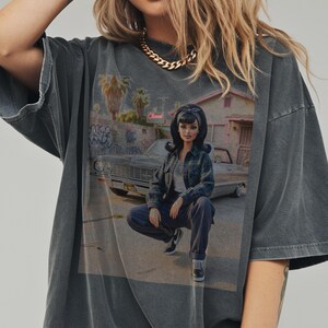 May include: Dark gray t-shirt featuring a graphic of a doll in a plaid shirt and jeans, squatting in front of a car and a building with graffiti. The shirt has a vintage, washed look.