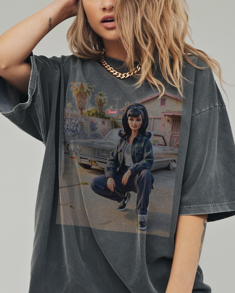 May include: Dark gray t-shirt featuring a graphic of a doll in a plaid shirt and jeans, squatting in front of a car and a building with graffiti. The shirt has a vintage, washed look.