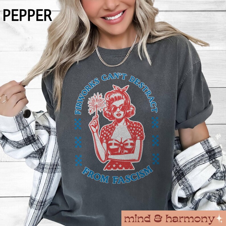 May include: Charcoal grey t-shirt featuring a retro graphic of a woman with sunglasses holding a firework. The text reads "Fireworks Can't Distract From Fascism" in blue and white. The shirt has a vintage, worn appearance.
