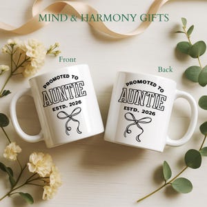 May include: Two white ceramic mugs with the text "PROMOTED TO AUNTIE ESTD. 2026" in black. Each mug has a bow graphic. The mugs are surrounded by flowers and greenery. The words "MIND & HARMONY GIFTS" are at the top.