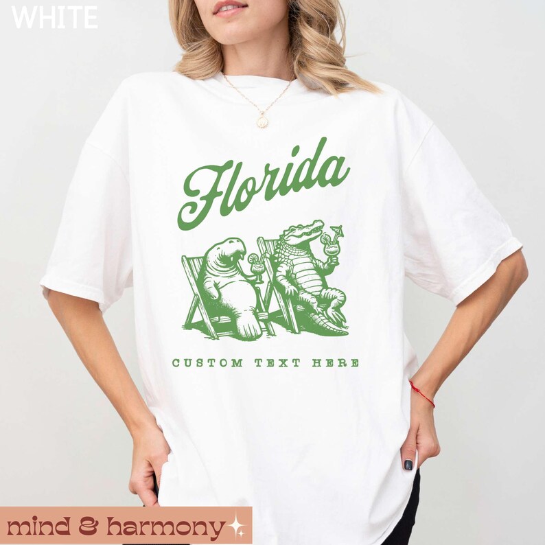 May include: White t-shirt with a green graphic of a manatee and alligator holding drinks. The text "Florida" is above the graphic and "Custom Text Here" is below.